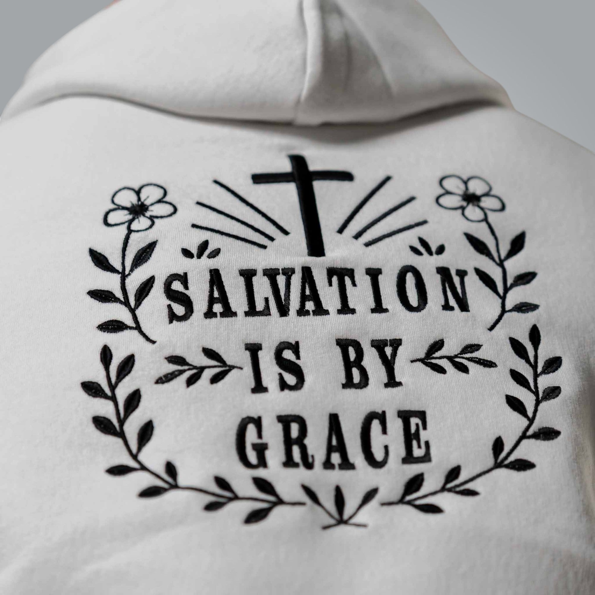 Close up of black embroidered Salvation Is by Grace text on the back of men’s white hoodie