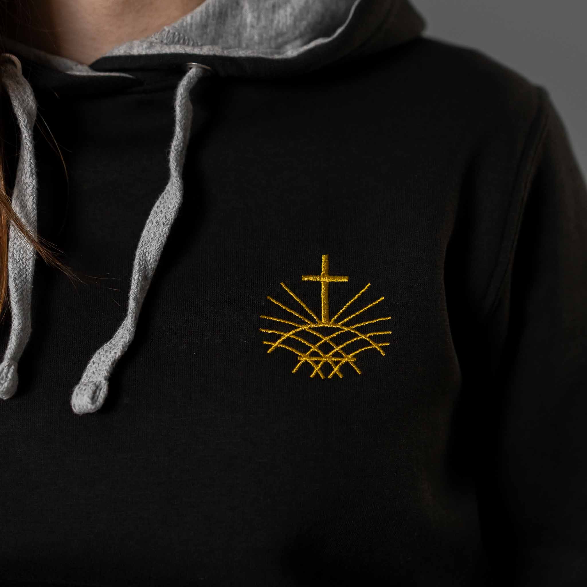 Close up of gold embroidered Loom in Light cross emblem on black women’s hoodie