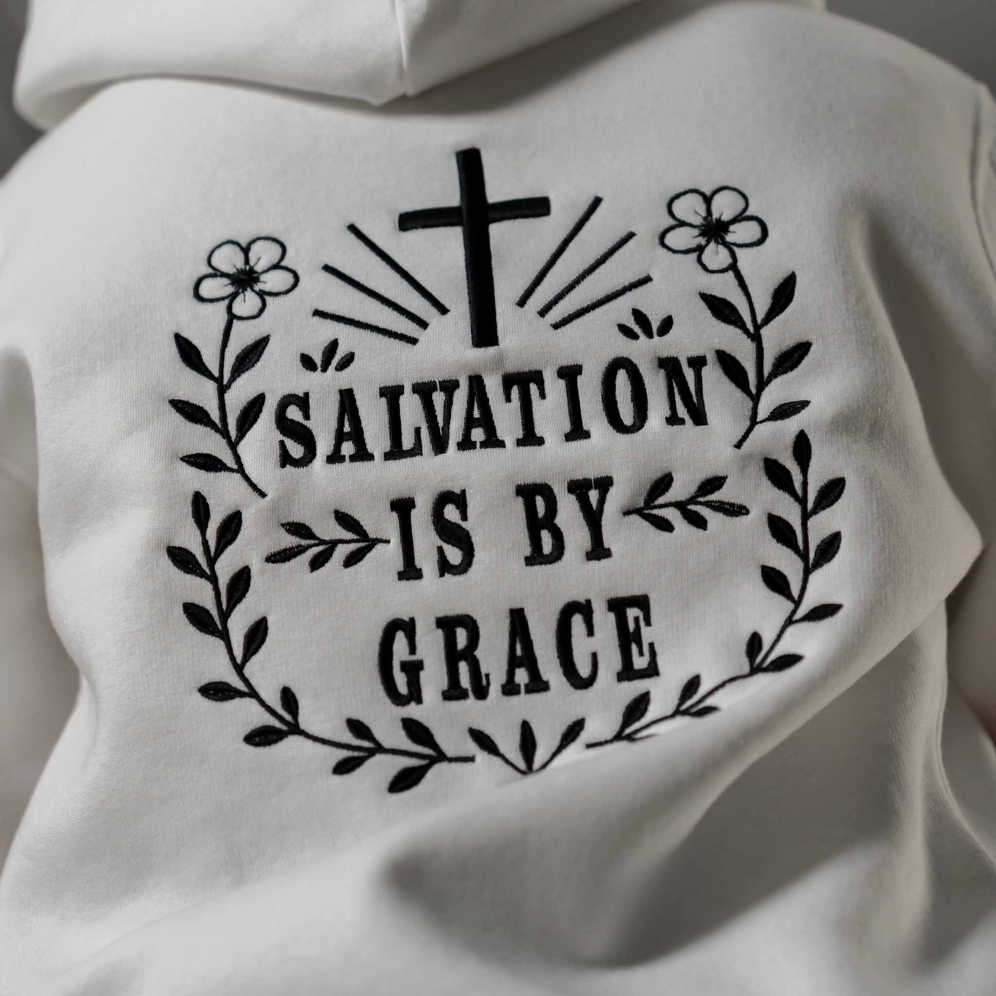 Close up of black embroidered Salvation Is by Grace text on white hoodie